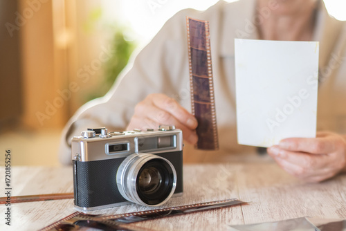 Person's hands holding an undeveloped film strip and a blank photographic print, with a vintage analog camera and more film strips on a wooden table, representing classic photography and nostalgia