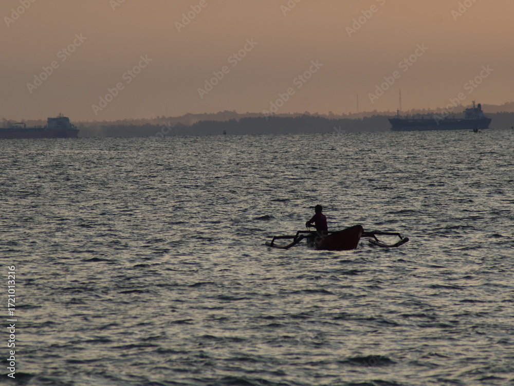 Fototapeta premium Silhouette of a traditional fisherman in a small outrigger boat on the open sea, with large cargo ships and a coastal skyline in the distant background at sunset.
