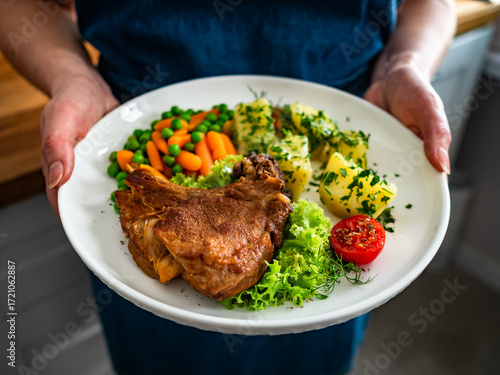 Woman holding plate with baked veal cutlet with bone, boiled potatoes, mini carrots and green peas, close-up