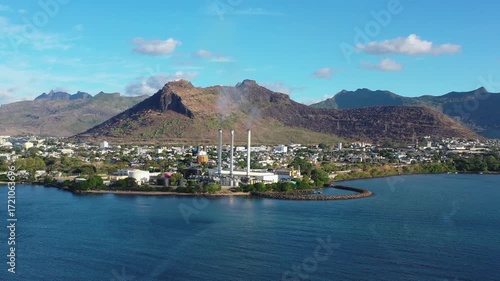 Aerial view of a power plant with tall smokestacks emitting smoke, nestled near mountains and the calm blue ocean, Port Louis, Port Louis District, Mauritius.