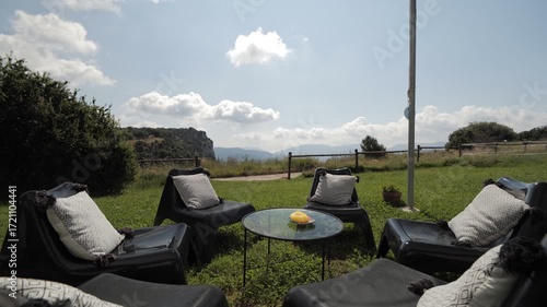 Outdoor seating area with black lounge chairs and a glass table. Scenic view of mountains and blue sky with clouds. Green grass surrounds the area.
