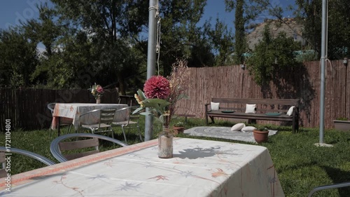 A sunny outdoor garden setting with a table and chairs. A vase with flowers is on the table. Green grass and trees surround the area, creating a relaxing atmosphere.