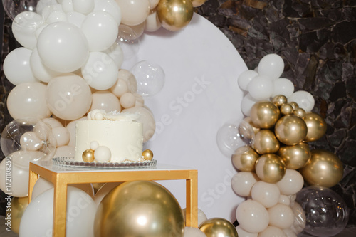 A chic and minimalist white cake decorated with golden spheres, showcased on a gold stand against a stylish photo zone with a white, beige, and gold balloon arch 