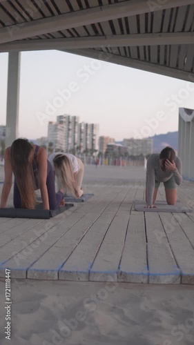 Pregnant women are practicing yoga together on the beach at sunrise. They are using yoga mats on a wooden deck, performing gentle poses for relaxation, strength, and mindfulness in a calm outdoor