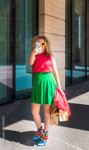 a beautiful young woman in bright clothes with a phone and bags takes selfies or takes pictures of the city on a walk. the concept of fashion, youth, mobile photography, tourism
