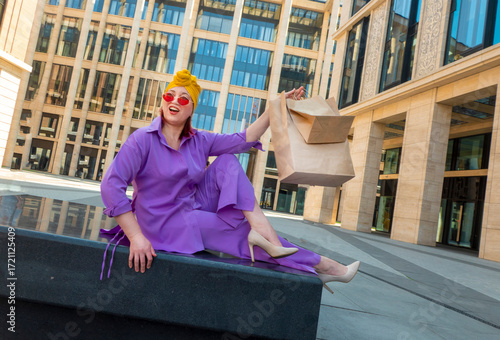 woman with craft shopping bags in the city. The concept of fashionable Consumerism and ecology. An adult Asian woman in a purple suit with red and yellow accents enjoys shopping . a sunny day.