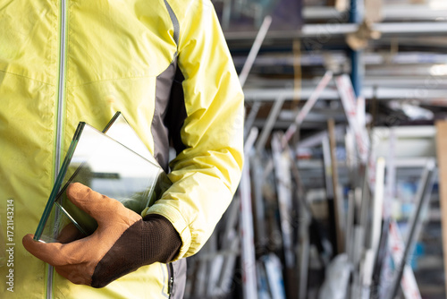 A glazier stands wearing gloves holding thick glass in a glass factory.