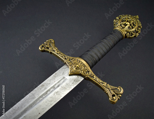 Ornate medieval broadsword with a beautifully crafted golden hilt, black wrapped grip, and a weathered steel blade on a dark background