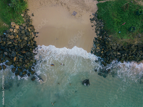 Aerial view of the meeting point of the ocean and the sandy beach, framed by rocky outcrops covered in green vegetation, Welligama, Sri Lanka.