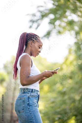 Young woman with braided ponytail smiling while interacting with smartphone outdoors in natural light