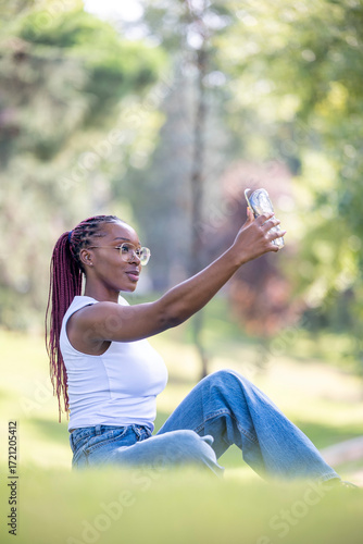 Young woman with braids taking selfie outdoors sitting on grass during sunny summer day