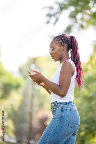 Stylish young woman typing on smartphone outdoors surrounded by greenery during sunny summer day