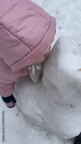 Little toddler girl in warm outerwear licking snow snowman outside in winter childhood