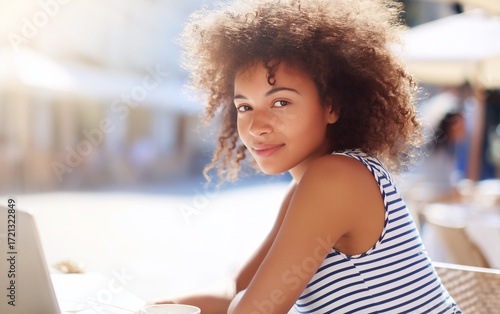 A beautiful smiling young Black woman working on laptop at a European street cafe on a warm sunny summer day.