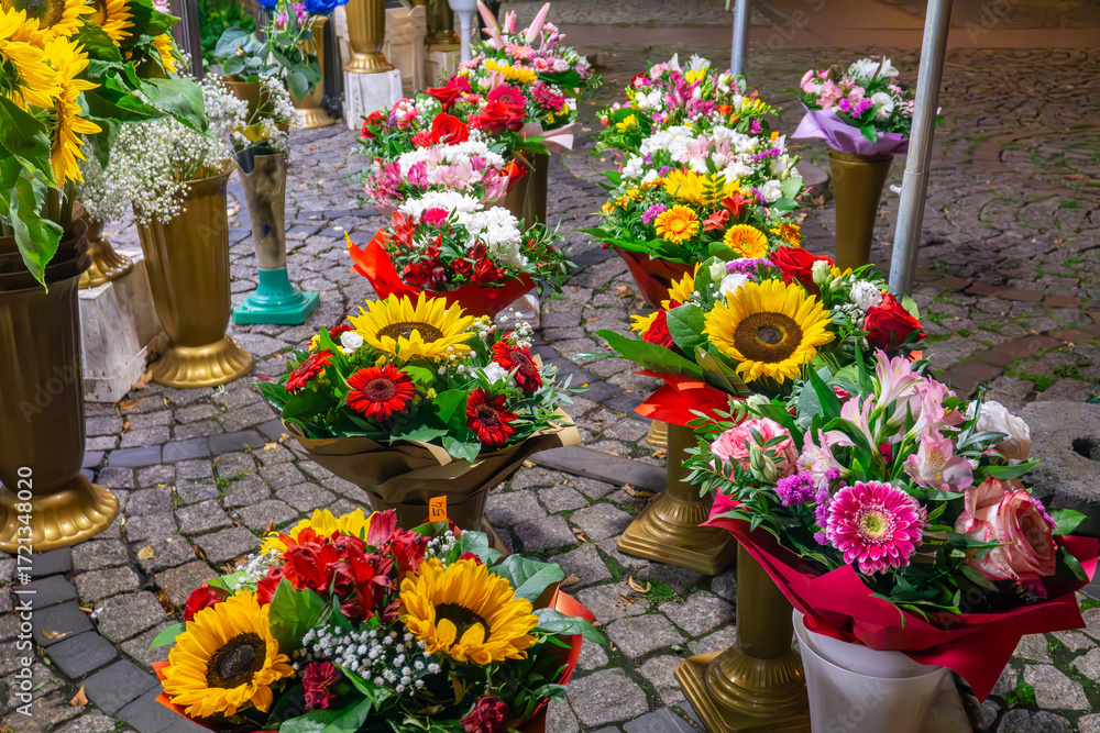 Obraz premium Flower bouquets with yellow sunflowers, roses and gerberas displayed in decorative stands on cobblestone street outside florist shop.