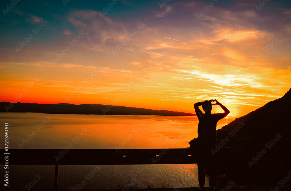 Fototapeta premium Silhouette of a person making a heart shape with hands at sunset, overlooking calm sea and mountains, warm sky colors creating romantic and peaceful mood