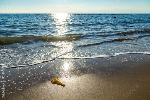Fototapeta Naklejka Na Ścianę i Meble -  Yellow toy rake left on sandy beach at the waterline, with gentle sea waves sparkling in sunlight. Summer holiday and childhood concept. Side, Antalya, Turkey.