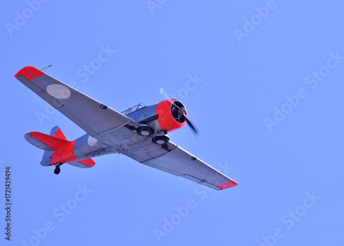 Photograph of a vintage aircraft, possibly a World War II military trainer or patrol plan, flying against a clear blue sky. The aircraft has red and silver accents, with a visible rotating propeller