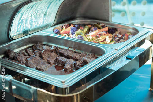 buffet chafing dish filled with juicy beef slices in rich gravy on the left and a colorful medley of roasted vegetables including carrots and purple cauliflower on the right.