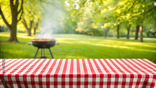 A checkered red and white tablecloth covers a picnic table in a park with a barbecue grill smoking in the background