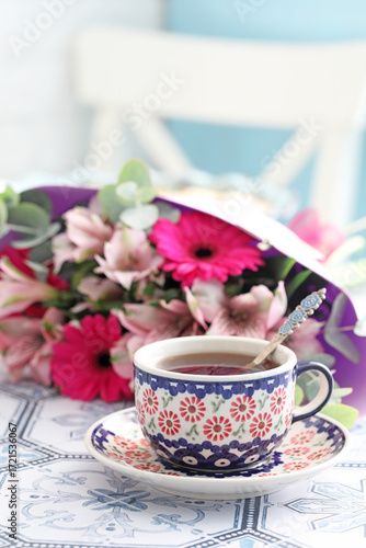 A beautiful ceramic cup and saucer with tea on a table. Flowers in the background. Selective focus.