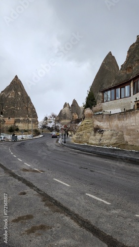 Winding road in Cappadocia, Turkey, flanked by distinctive fairy chimneys—tall, thin rock formations. The landscape is rugged, with layers of sedimentary rock creating unique textures and colors in sh