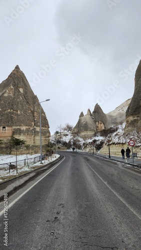 Winding road in Cappadocia, Turkey, flanked by distinctive fairy chimneys—tall, thin rock formations. The landscape is rugged, with layers of sedimentary rock creating unique textures and colors in sh