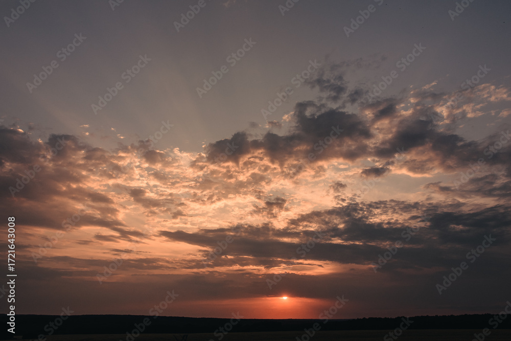 Fototapeta premium A picturesque sunset scene featuring a vibrant array of colors in the sky. The sun is setting on the horizon, casting a warm glow on the clouds and the land below.