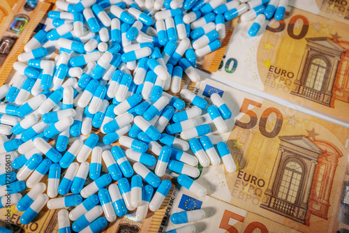 Blue and white capsules scattered over 50 euro banknotes, symbolizing pharmaceutical costs, healthcare expenses, medical economy, and drug industry.