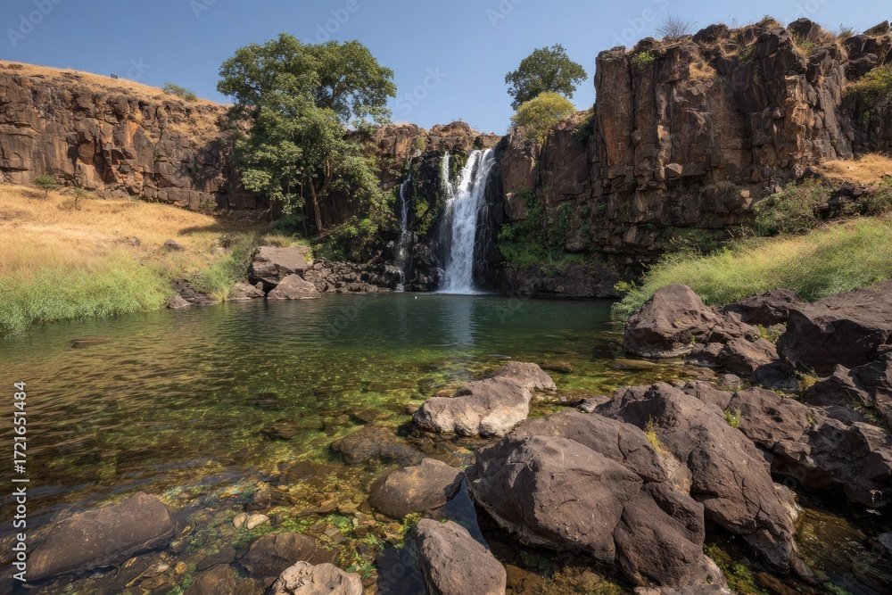 Obraz premium A tranquil waterfall cascades into a crystal-clear pool surrounded by dark, rugged rocks and lush greenery.