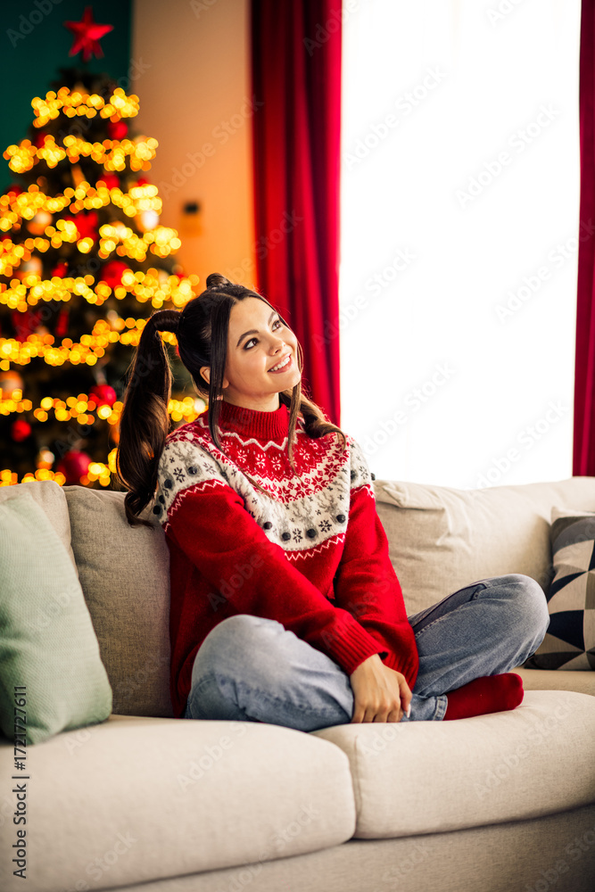 Fototapeta premium Festive woman on cozy sofa embracing holiday cheer with Christmas tree lights, ornament-filled background, and joyful atmosphere