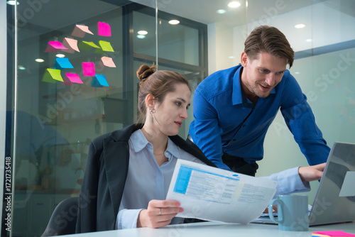 Efficient teamwork in modern office setting showcases woman reviewing documents while man assists her with laptop. Colorful sticky notes in background add creative touch