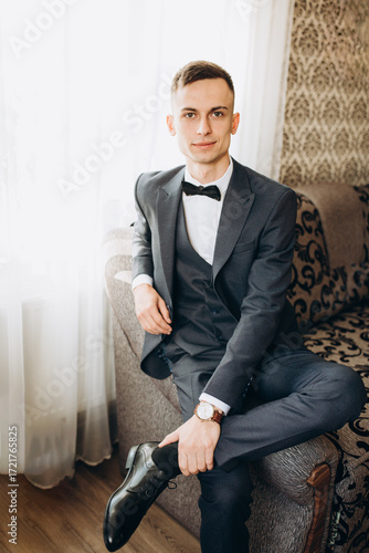 Groom in stylish dark suit with bow tie sitting indoors on a sofa. Young man with classic watch and polished shoes, posing confidently on his wedding day. Elegant portrait in formal attire.