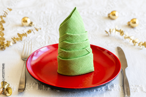 Green linen napkin folded in shape of Christmas tree and knife, fork on white tablecloth with golden christmas decoration. New Year table setting. 
