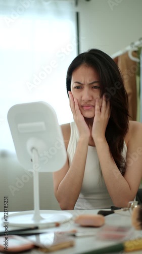 Vertical video. Worried Asian woman looking at mirror, touching dry irritated skin and acne spots. Female feeling unhappy with wrinkles and face dehydration, searching for skincare solution. 
