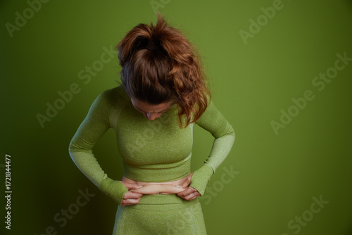 A 30-something woman with red curly hair and athletic wear looks down as she pinches skin on her lower abdomen in a softly lit studio.