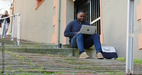 Placing laptop on lap, African American man typing on steps, sipping travel mug and studying