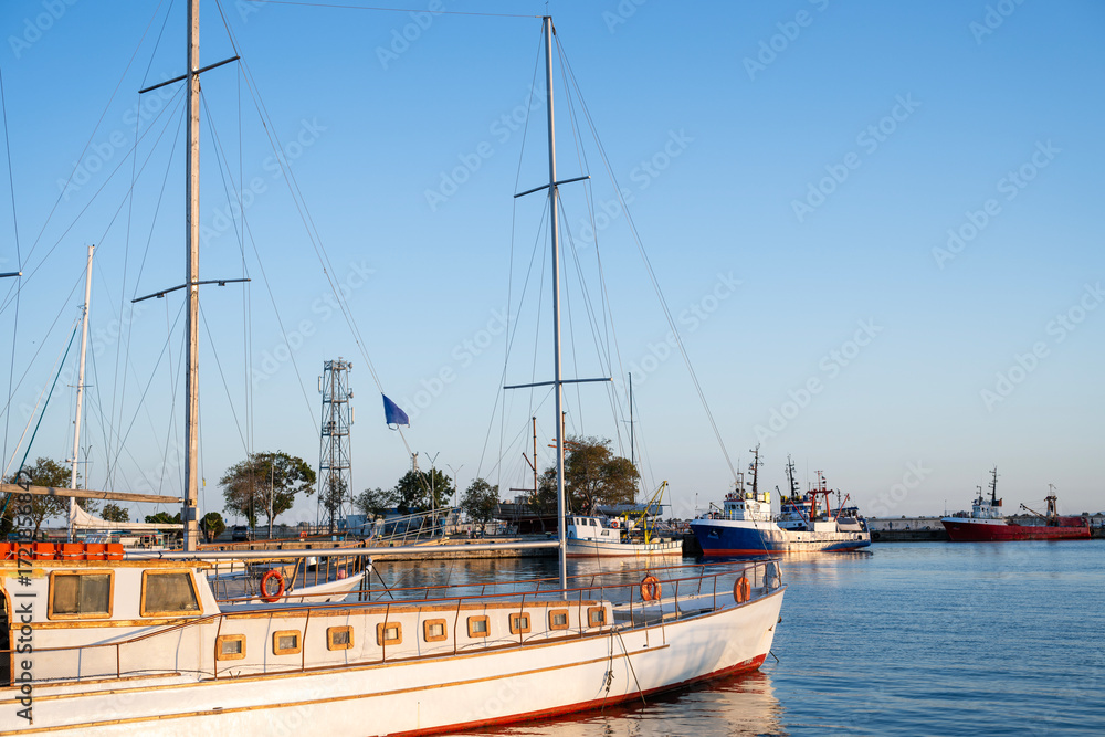 Fototapeta premium A wooden sailboat with white hull rests in a calm harbor, surrounded by colorful ships and reflected in tranquil water.