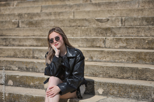 Canvas Print Young, beautiful, blonde woman, with leather jacket, snake miniskirt and sunglasses, posing looking at the camera sitting on the stairs, relaxed and calm