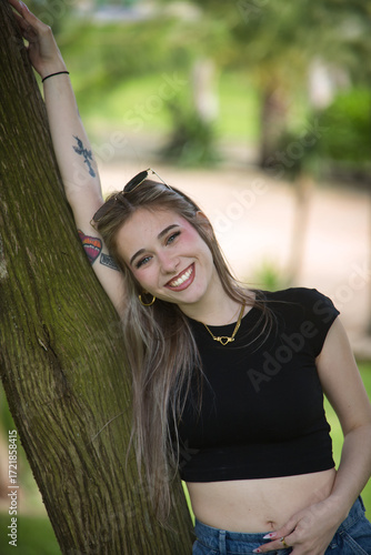 Portrait of a young woman, beautiful, blonde, green eyes, wearing a black t-shirt, looking at the camera smiling and happy leaning against the trunk of a tree. Concept of smile, teeth, happiness.