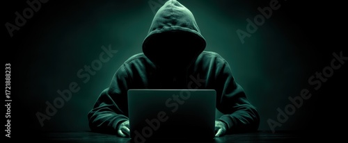 The hooded figure using a laptop in a darkened environment.