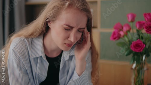 Young woman with long blonde hair, sitting on a sofa in her living room, is suffering from a severe headache, massaging her temples with a pained expression and closed eyes
