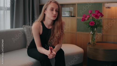 Young woman sitting alone on a sofa in a modern living room, pensive and overwhelmed by heartbreak, loneliness and anxiety, conveying sadness, stress and emotional exhaustion