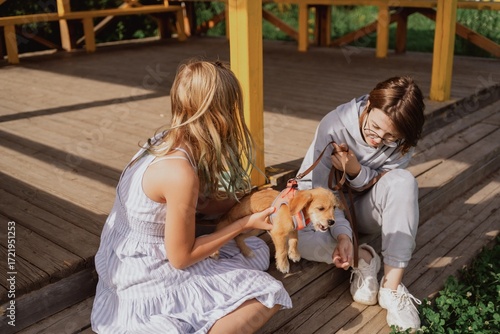 A young woman and her teenage daughter walk their playful puppy in a sunny garden, sharing a multigenerational moment of pet care and family bonding. Authentic, unaltered photography