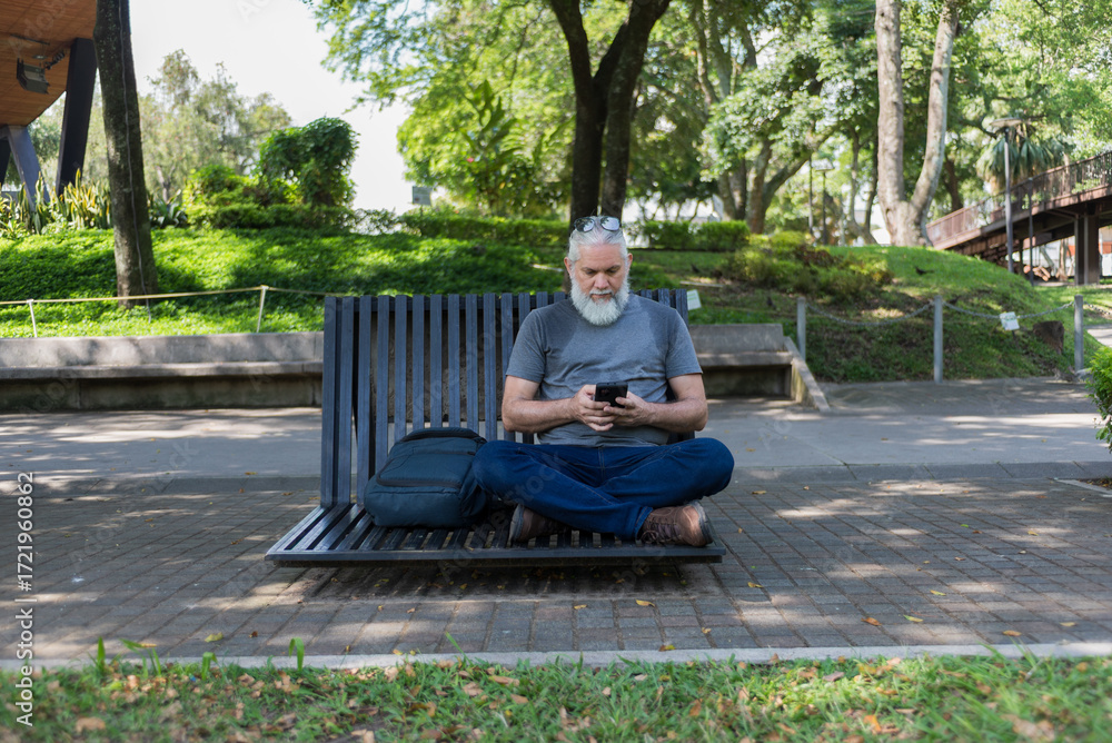 Obraz premium An older man with a gray beard sits cross-legged on a modern park bench, using his smartphone.