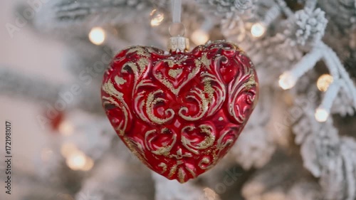 Christmas ornament red heart-shaped with golden decorative patterns close-up, hanging on Christmas tree. Warm lights illuminate festive scene, adding romantic cozy holiday feel. Winter family holiday
