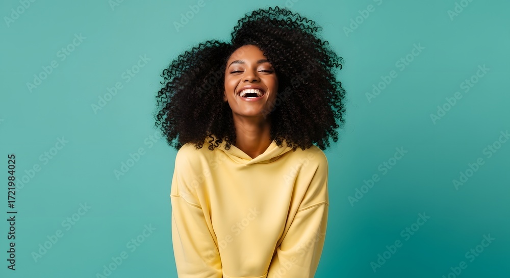 Fototapeta premium An energetic woman with voluminous afro hair and a bright smile expresses pure joy. Her yellow hoodie and the vibrant teal background create a dynamic and cheerful atmosphere.