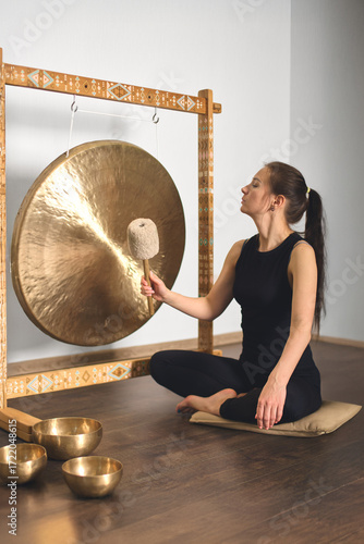 Wallpaper Mural Woman seated in serene studio striking large gong with mallet. Surrounded by singing bowls on dark wooden floor. Soft lighting casts calming ambiance Torontodigital.ca