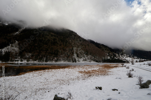 Obraz na płótnie Campo nevado junto a una lámina de agua tranquila, con laderas boscosas cubiertas de niebla bajo un cielo gris con claros