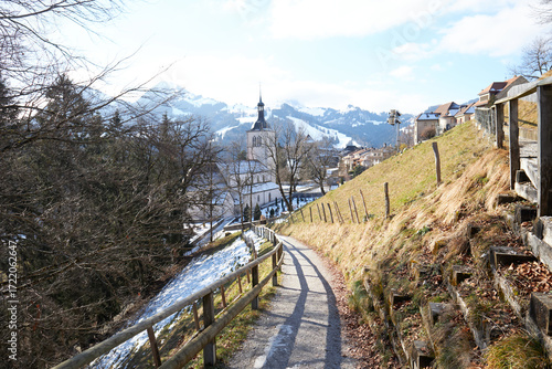 Tableau sur toile Sendero asfaltado entre vallas y árboles desnudos, descendiendo hacia iglesia y casas con montañas nevadas al fondo